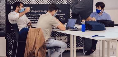 Three men, two wearing masks, are engaged in a discussion at a table with laptops in an office setting.