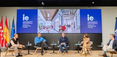 A panel discussion at IE University School of Architecture & Design, featuring five individuals seated in front of banners with university and national flags.