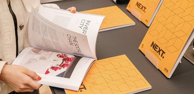 A person is flipping through a magazine on a table while there are additional magazines displayed in the background.