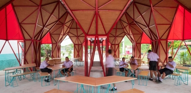 A modern angular canopy structure with red fabric and wooden tables, populated by students interacting in a lively environment.