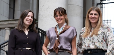 Three women are smiling and posing together outdoors near a building.