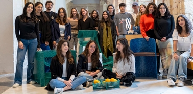 A group of young adults posing together in a modern indoor space, some seated on blue chairs, others standing, with visible smiles.