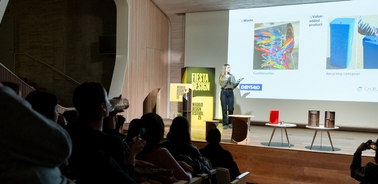 A speaker presents on stage at a design event while an audience listens attentively.
