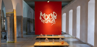 A modern art exhibition in a gallery with a vibrant red backdrop featuring white text and several art installations on display tables.