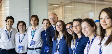 A group of young adults and a man in formal attire are smiling together in a well-lit indoor setting.
