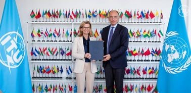A formal agreement signing ceremony is taking place between two officials in front of a backdrop of flags.