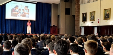 A speaker presents to a large audience of students in a school assembly hall, with a projected image behind him.