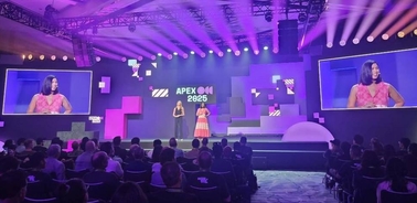 A conference scene with two speakers on stage in front of a large audience and screens displaying the event logo.