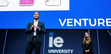 A man stands speaking at a podium with 'VENTURE' displayed on a large screen, and a woman is seated beside him, both at an event for IE University.