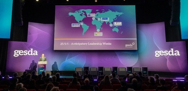 A person is giving a presentation at a conference with a world map and event dates displayed on the screen behind them.