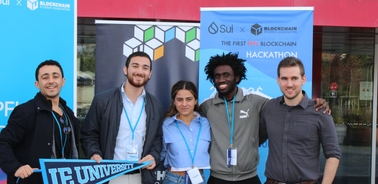 A group of five people smiling in front of a banner at a blockchain hackathon event.