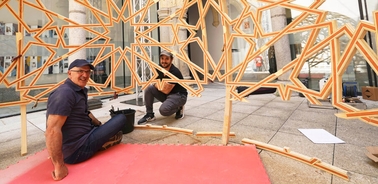 Two men sitting on a red carpet, installing a large geometric sculpture in an outdoor setting.