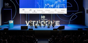 A panel of executives participating in a conference discussion on a stage sponsored by multiple companies.