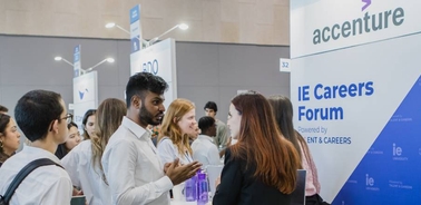 A group of people engaging in conversation at a career forum with Accenture and IE University banners in the background.