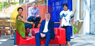 A group of four diverse people smiling and sitting on a colorful public bench in a sunny urban setting.