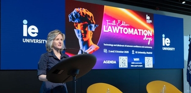 A woman stands at a podium in front of a large screen displaying event information and a sculpture.