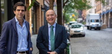 Two men in business attire walking on a city street with one looking at the camera and the other looking away.