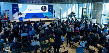 A speaker presents at a podium in front of an audience in a conference room with large windows and banners.