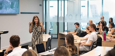 A woman in a zebra-striped dress is speaking in front of an attentive audience in a modern classroom.