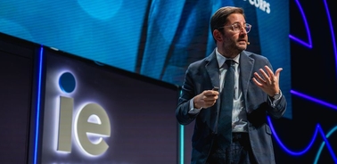 A speaker is presenting on stage at a conference, gesturing while holding a microphone.