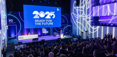 The image depicts a graduation ceremony at IE University featuring a large digital screen displaying '2025 READY FOR THE FUTURE'.