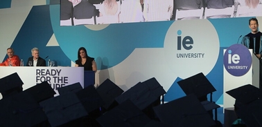 A graduation ceremony at IE University with speakers on stage and empty chairs in the foreground.