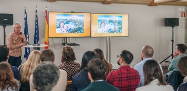 A man presents with two large screens showing graphics during a conference in a room filled with seated attendees.