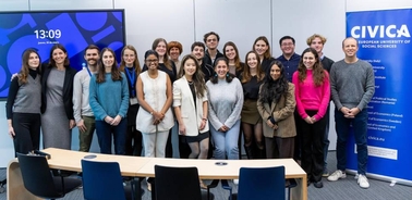 A group of students and professionals standing in a meeting room with a digital clock and CIVICA banner.