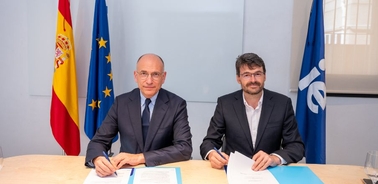 Two men are sitting at a table signing documents with flags in the background.