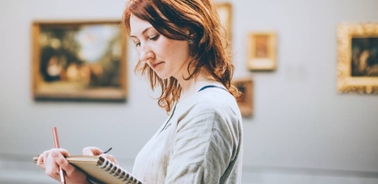 A woman sketches in a gallery surrounded by paintings.
