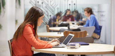 A woman works on a laptop at a café-style table while others are engaged in discussion in the background.
