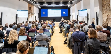 A large audience is attending a presentation in a modern conference room.