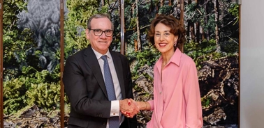 A man and a woman in business attire shaking hands in front of a wall with nature-themed artwork.