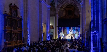 An event with attendees seated in a cathedral listening to a speaker on stage illuminated by blue lights.