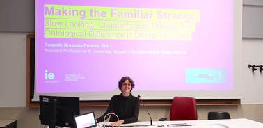 A woman sitting at a desk in front of a presentation screen with the title of a lecture.