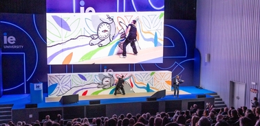 Two performers are engaging in a playful mock fight on stage in front of a large audience with a colorful backdrop.