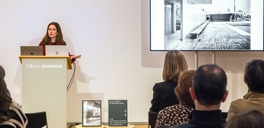 A woman is giving a presentation with a large image displayed on a screen behind her.