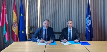 Two men are sitting at a table, signing documents, with flags in the background.