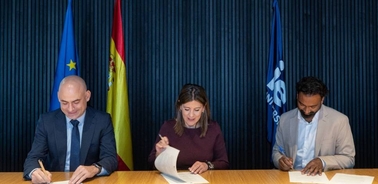 Three individuals are signing documents at a table with flags in the background.