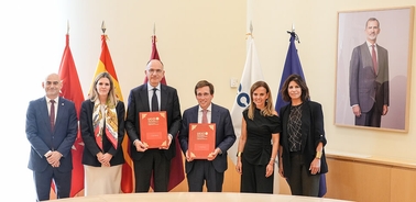 A group of officials standing together in an office, holding documents with flags in the background.