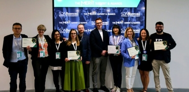 A group of people standing on stage, each holding a certificate, with banners related to 'The MERIT Summit & Awards' in the background.