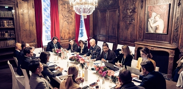 A formal meeting taking place in an elegant room with a long table and attendees discussing.