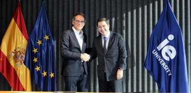 Two men in suits shaking hands in front of flags of Spain, the European Union, and IE University.