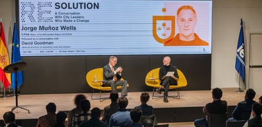 Two men are engaged in conversation on stage in front of an audience, with banners displaying city flags and names.