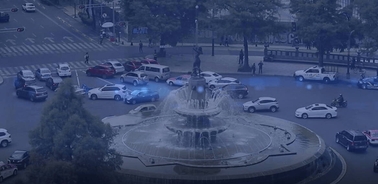 Aerial view of a busy city intersection with a circular fountain in the center surrounded by various vehicles and pedestrians.