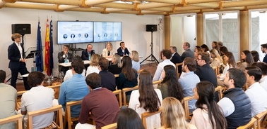 A speaker presents at a conference in front of an audience in a room with banners and a logo.