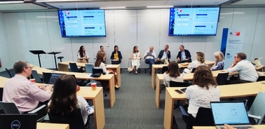 A group of professionals sitting in a seminar room with presentations displayed on screens.