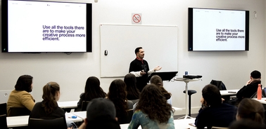 A speaker presents to an audience in a classroom setting with two screens displaying a quote.