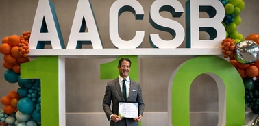 A man is holding a certificate in front of a colorful backdrop celebrating AACSB's 110th anniversary.