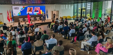 A conference setting with attendees seated observing a presentation at a brightly lit venue with prominent displays showing LGBT+ flags and supportive messages.